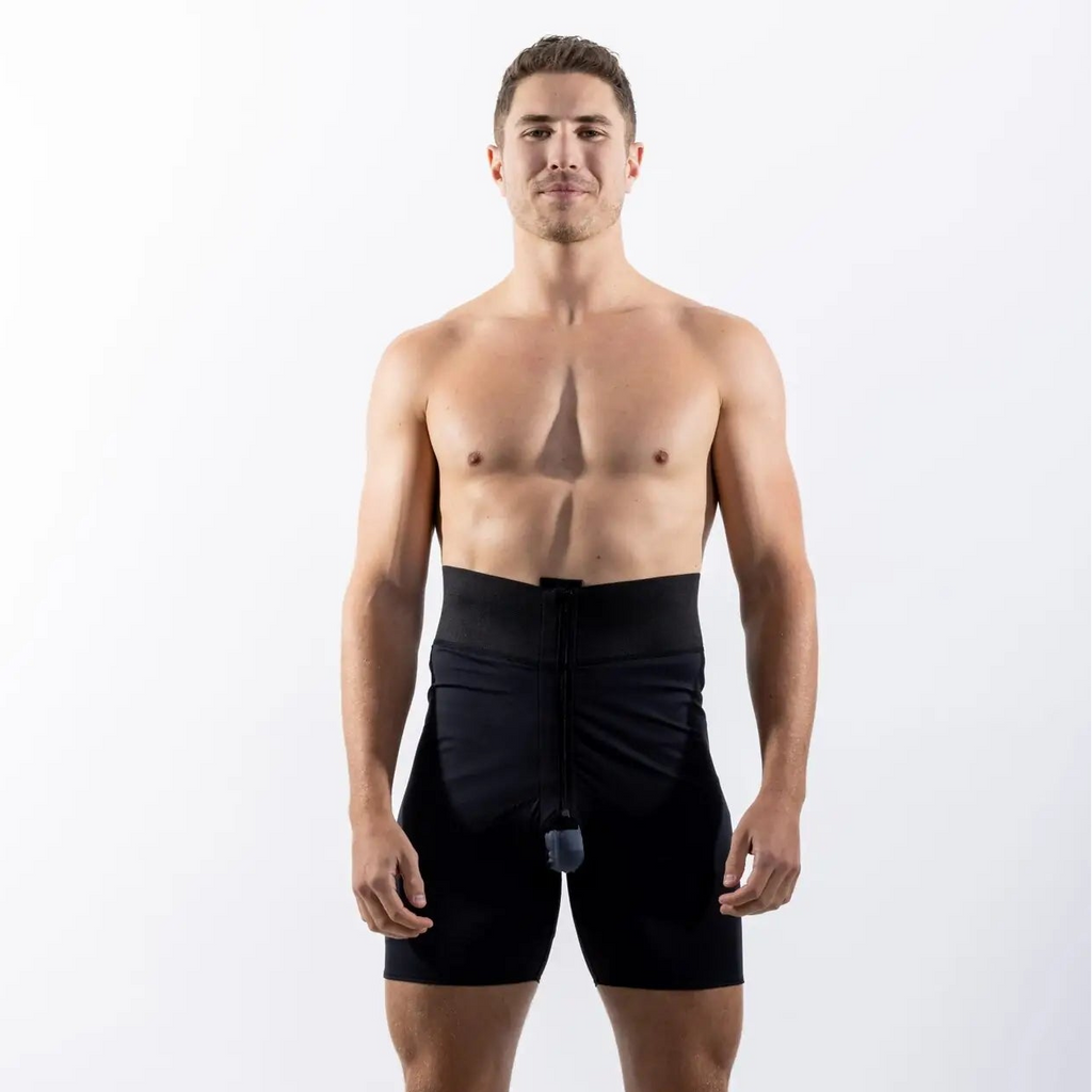 A shirtless man standing against a white studio background wearing black athletic shorts with a small attached device near the waist.