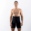 A shirtless man standing against a white studio background wearing black athletic shorts with a small attached device near the waist.