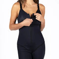 A woman modeling a black shapewear bodysuit with shoulder straps, a front zipper, and built-in shorts with lace trim at the hem, set against a plain white background.