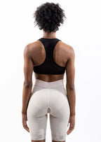Rear view of a model wearing a black sports bra and beige high-waisted compression shorts against a white studio background.