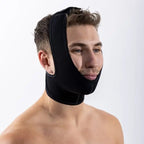 A shirtless man wearing a black neoprene chin and jaw support brace against a plain white studio background.