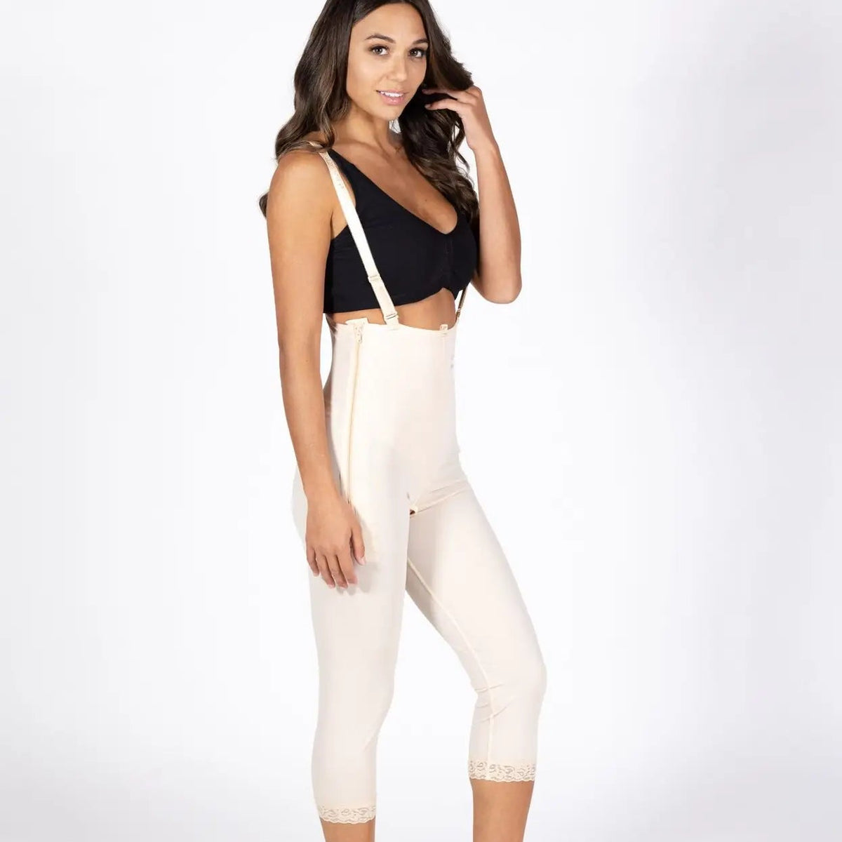 A woman modeling high-waisted, mid-thigh compression or shaping pants in a light cream color, paired with a black crop top, against a plain white studio background.